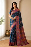 A woman modeling a blue patterned saree with a rich magenta and gold border, standing in a warm, neutral-toned indoor setting.